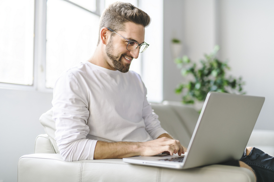Young attractive guy is browsing at his laptop, sitting at home on the cozy beige sofa at home, wearing casual outfit