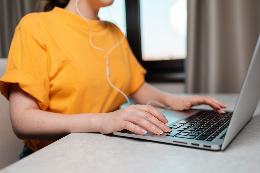A woman is sitting at a desk with headphones on, typing on a laptop. Hands close-up. The concept of online work and education.
