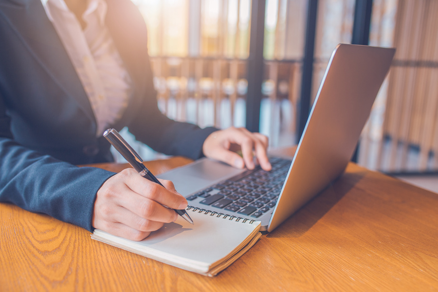 Business women are taking notes on paper with a black pen, and she is using a laptop computer on a wooden desk in the office.