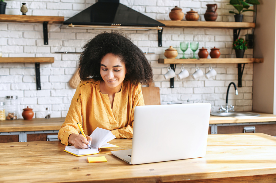 A beautiful black woman in smart casual wear is using laptop computer in the kitchen, a thoughtful female employee is writing in planner. Work remotely from home