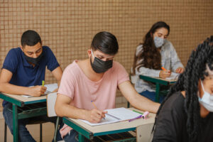 Latin students in the classroom. Male student wearing mask and writing in notebook with a pen. Covid-19. Pandemic.