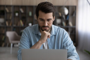 Thoughtful young male manager in eyeglasses looking at laptop screen, considering problem solution, preparing research report, analyzing sales statistic, developing strategy, working on online project