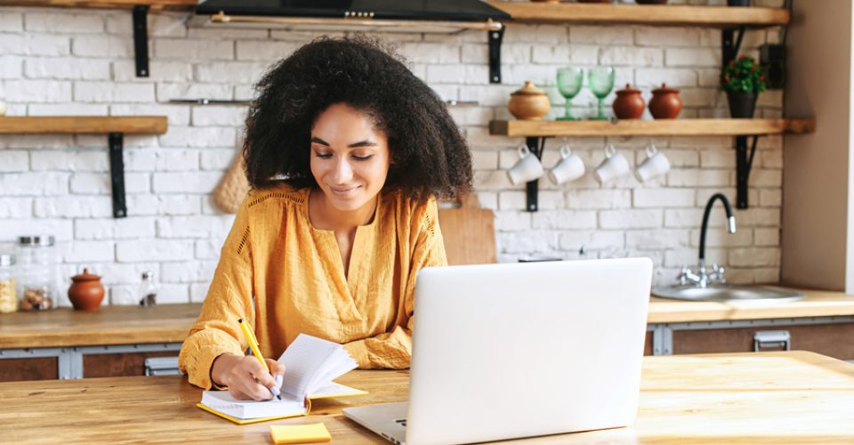 A beautiful black woman in smart casual wear is using laptop computer in the kitchen, a thoughtful female employee is writing in planner. Work remotely from home