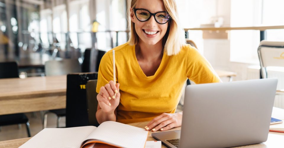 Photo of beautiful happy woman looking at camera while working and sitting at table in open-plan office