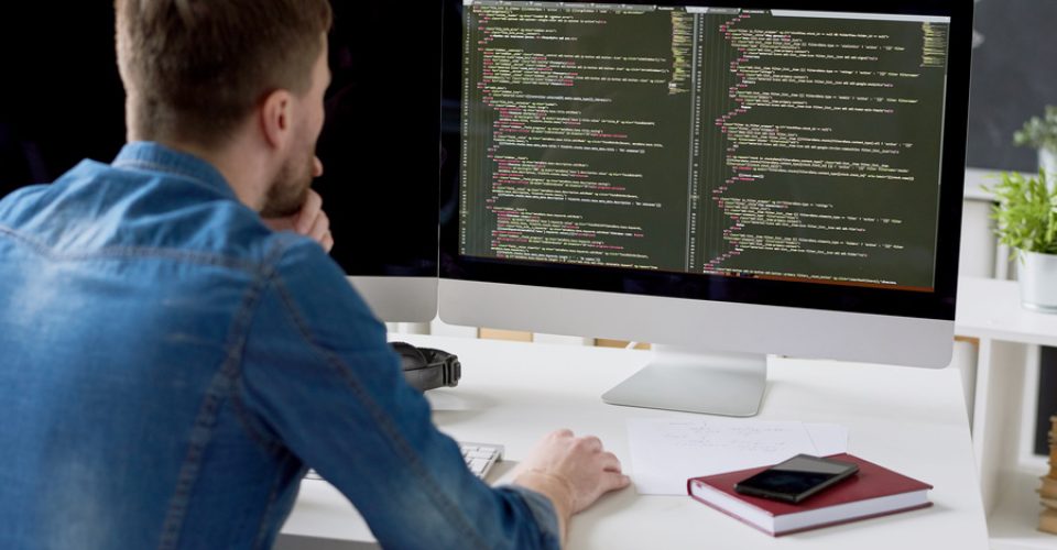 Rear view of concentrated thoughtful male programmer viewing computer language code on computer monitor while working on new program in modern office