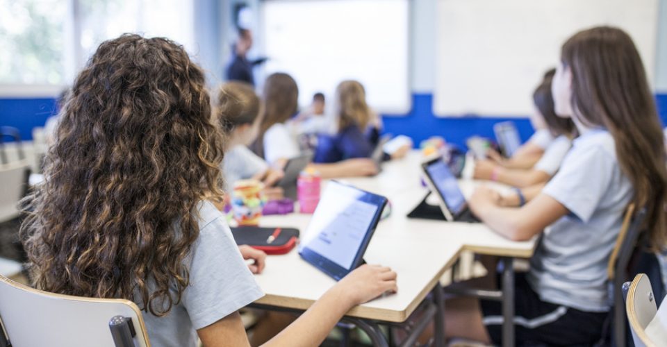 girl with brown curly hair studies in class with her tablet next to her classmates while they listen to the teacher