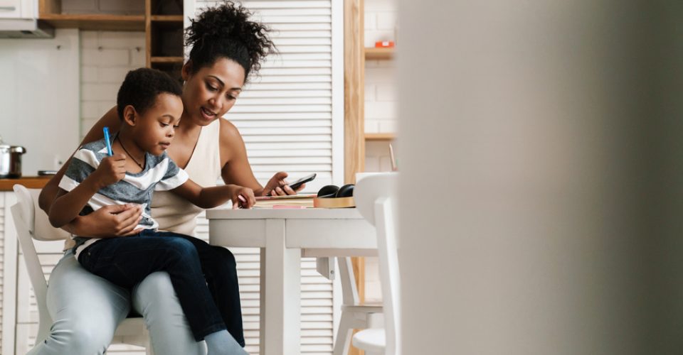 Black mother and son writing and using mobile phone while sitting together at home