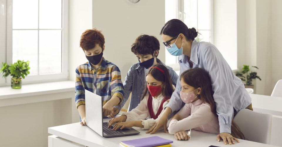 Primary school children and teacher using laptop in classroom. Happy junior students and tutor wearing medical face masks coding video game. Computer lab, technology, basic programming, STEM education