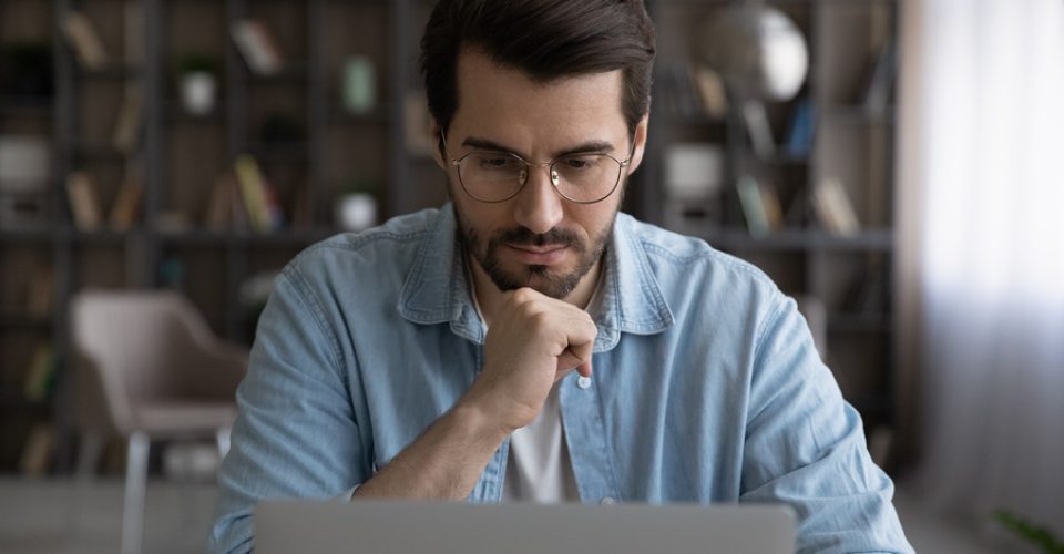 Thoughtful young male manager in eyeglasses looking at laptop screen, considering problem solution, preparing research report, analyzing sales statistic, developing strategy, working on online project