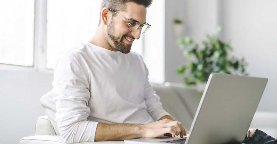 Young attractive guy is browsing at his laptop, sitting at home on the cozy beige sofa at home, wearing casual outfit