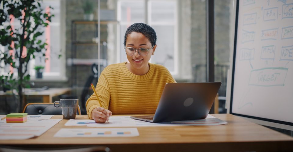 Portrait of Young Latin Marketing Specialist in Glasses Working on Laptop Computer in Busy Creative Office Environment. Beautiful Diverse Multiethnic Female Project Manager is Browsing Internet.