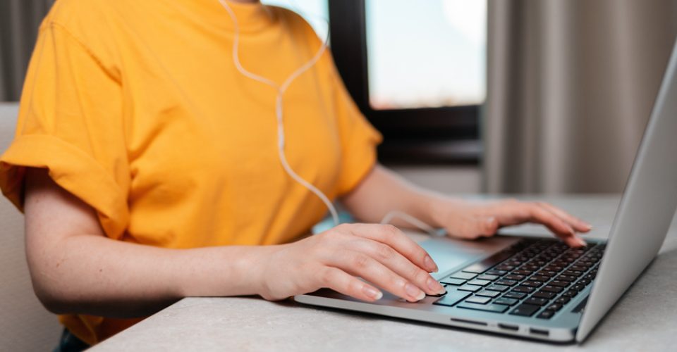 A woman is sitting at a desk with headphones on, typing on a laptop. Hands close-up. The concept of online work and education.