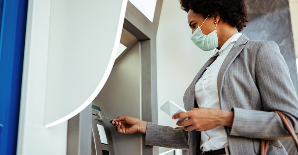 Low angle view of African American businesswoman inserting credit card and withdrawing cash at ATM while wearing protective mask on her face.
