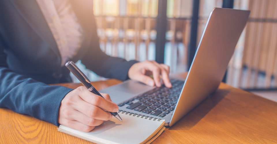 Business women are taking notes on paper with a black pen, and she is using a laptop computer on a wooden desk in the office.