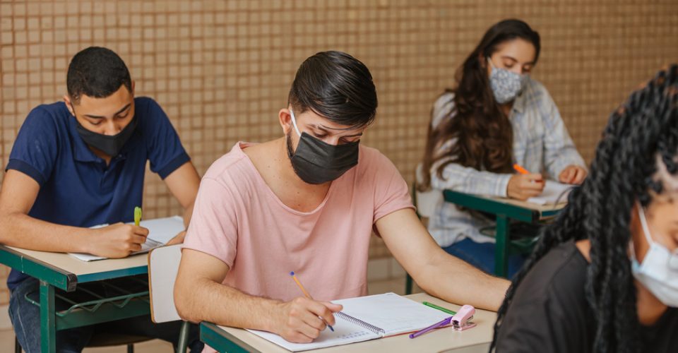 Latin students in the classroom. Male student wearing mask and writing in notebook with a pen. Covid-19. Pandemic.